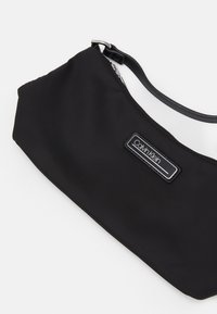 Sac d'épaule en nylon noir avec une fermeture éclair, présentant une texture lisse et une étiquette rectangulaire du logo Calvin Klein à l'avant.