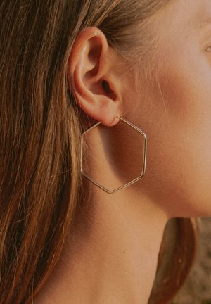 Goldfarbene sechseckige Creole, dünnes Drahtdesign, verfügt über eine polierte Oberfläche, sicher durch das Ohrläppchen befestigt, wodurch die Kontur des Ohrs betont wird.