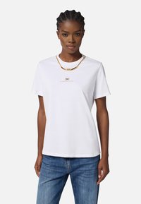 White cotton t-shirt with short sleeves, featuring a gold logo and a gold chain necklace layered on top. Model wearing blue jeans.
