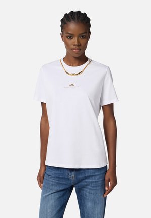 White cotton t-shirt with short sleeves, featuring a gold logo and a gold chain necklace layered on top. Model wearing blue jeans.