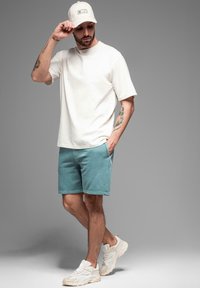 Bílý tričko s krátkým rukávem, tyrkysové šortky a béžové sportovní boty. Doplňky zahrnují čepici světlé barvy. Minimalistický design a nenucený vzhled.