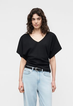 Kvinne med krøllete hår iført en svart V-hals kortermet bluse og lyseblå jeans med svart belte, stående mot en ensfarget bakgrunn.