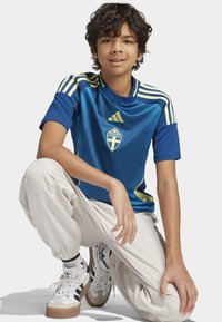 Modrý sportovní dres se žlutými akcenty a emblémem "SVERIGE"; spárovaný se světlými kalhotami a bílými teniskami s černými pruhy.