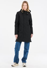 Parka noir imperméable avec capuche, fermeture éclair et cordons ajustables. Possède une texture lisse et une coupe mi-longue. Associée à un jean bleu.