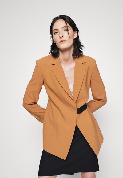 Blazer marron ajusté avec des épaules structurées, des revers en pointe et un seul bouton. Porté sur une jupe noire, avec une ceinture noire cintrée.