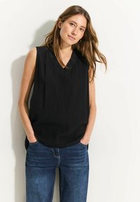 Zwarte mouwloze top met een V-hals en een textuurstof, gecombineerd met blauwe wijde jeans. De top heeft een losse pasvorm en zijsplitten.