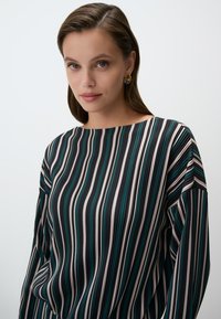 Blusa a righe verde, crema e nera. Presenta un ampia scollatura e un design fluido, con pieghe delicate e una texture di tessuto morbida.
