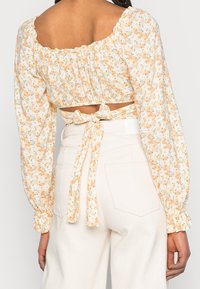 Blommig kortärmad blus i ljusgul med sammetsärmar, knytning i ryggen och små mönster av orangea och blå blommor på mjukt tyg.