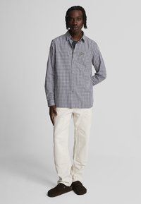 Chemise à carreaux bleue et blanche, en tissu léger. Associée à un pantalon crème et des mocassins marron foncé.