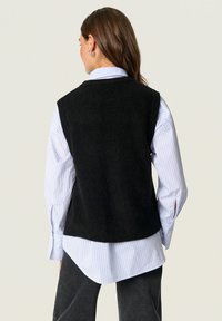Zwarte, geribbelde mouwloze trui gedragen over een blauw-witte gestreepte blouse, gecombineerd met donkergrijze broek.