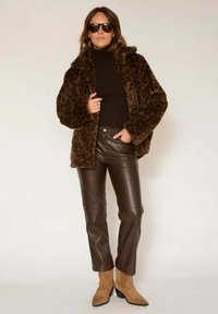 Veste en fausse fourrure à imprimé léopard, col roulé marron, pantalon en cuir marron et bottines beige, avec de grandes lunettes de soleil noires.