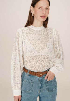 Blouse en dentelle blanche à manches longues et col haut, présentant un motif floral et des détails froncés, assortie à un jean bleu taille haute et une ceinture marron.