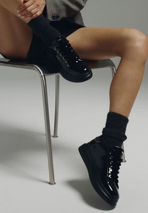 Zapatillas de charol negras con ojales dorados, cordones gruesos negros y una suela gruesa. Combinadas con calcetines oscuros texturizados y situadas frente a un fondo minimalista.