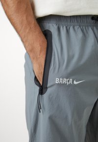 Pelēki sporta šorti, izgatavoti no viegla materiāla, ar rāvējslēdzēja kabatu un baltiem izšūtiem "BARÇA" un Nike logo.