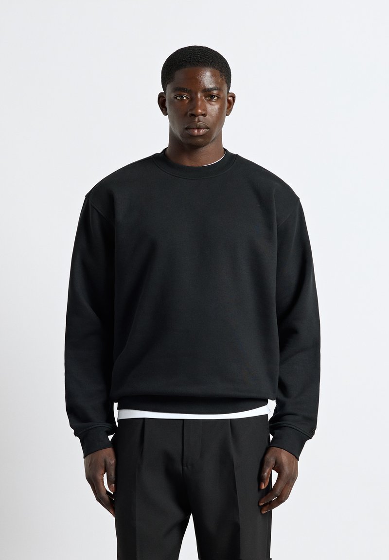 Junger Mann mit kurzen Haaren, trägt einen schlichten schwarzen Pullover über einem weißen Hemd und schwarzen Hosen, steht vor einem einfarbigen weißen Hintergrund.