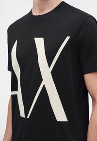Personne portant un t-shirt noir à manches courtes avec de grandes lettres blanches qui se chevauchent "A" et "X" imprimées sur le devant.