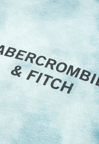 Tejido azul claro con un patrón de tie-dye, con un texto en negrita de color negro que dice "ABERCROMBIE & FITCH." Textura suave, diseño casual.