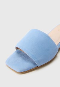 Infradito in suede blu con una fascia ampia e design a punta quadrata. La suola è piatta di un colore beige chiaro. Texture morbida in tutto il prodotto.