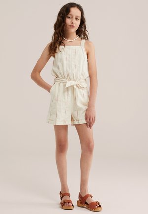 Niña con cabello largo y ondulado que lleva un mono sin mangas color crema con cinturón, sandalias marrones y un collar blanco de cuentas, situada frente a un fondo liso.