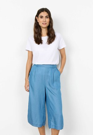 Frau mit langen dunklen Haaren, die ein weißes kurzärmeliges Hemd und lockere hellblaue Culottes trägt, steht mit einer Hand in der Tasche.