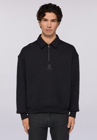 Svart långärmad tröja med dragkedja i kragen, ribbade muddar och nederkant, med en liten logotyp på bröstet. Tillverkad av mjukt tyg.