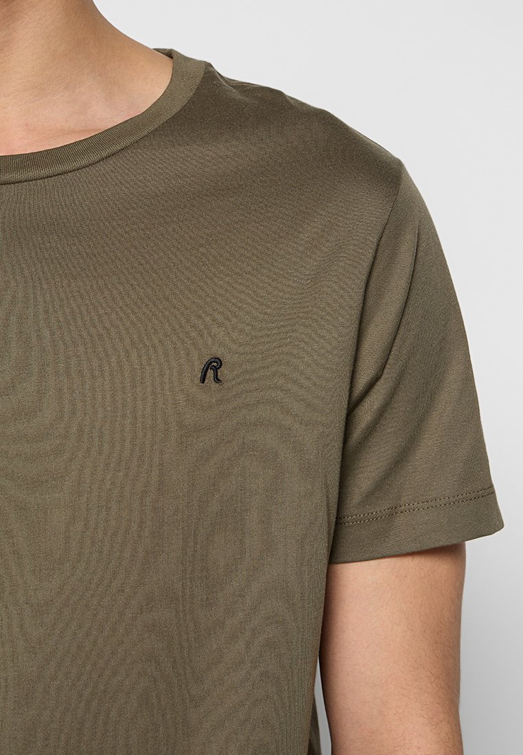 Nahaufnahme einer Person, die ein olivgrünes T-Shirt mit kurzen Ärmeln trägt, das auf der linken Brust ein kleines schwarzes, gesticktes "R" hat.