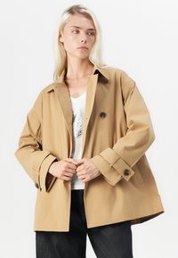 Trench coat bej dintr-un material durabil, având o croială lejeră, mâneci largi, prindere cu butoane mari și închidere cu un singur buton.