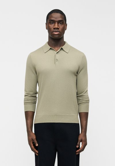Hombre vistiendo una camisa polo de manga larga beige claro y pantalones negros, de pie contra un fondo blanco liso.