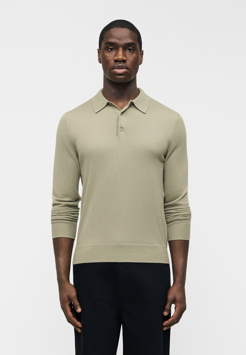 Homme portant un polo à manches longues beige clair et un pantalon noir, debout devant un fond blanc uni.