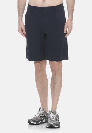 Uomo che indossa pantaloncini neri alla lunghezza del ginocchio e scarpe da corsa New Balance grigie, in piedi con le mani rilassate ai lati.