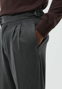 Grijze geplooide broek met een gladde textuur en zijzakken, voorzien van een riem met een rechthoekige metalen gesp. Donkerbruine mouw zichtbaar.