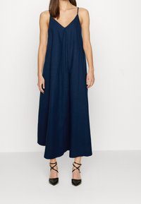 Mujer vestida con un vestido midi sin mangas de color azul marino con escote en V y tacones negros de punta con tiras alrededor del tobillo, de pie contra un fondo blanco.