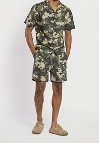 Man wearing green and beige camouflage short-sleeve button-up shirt with matching shorts and tan slip-on shoes, hands in pockets.
