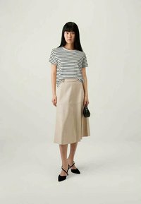 Nainen, jolla on päällään mustavalkoraidallinen t-paita, beige midi-hame, mustat kärkitossut ja joka kantaa pientä mustaa käsilaukkua.