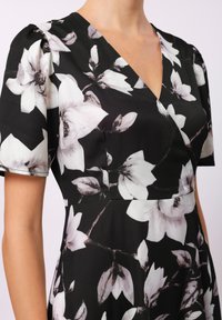 Svart kjole med hvit blomstertrykk, korte ermer, v-hals og strukturert stoff med en flytende design. Utsmykket med synlige blomsterdetaljer.