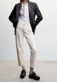 Blazer en cuir noir, chemise blanche à boutons, jeans droits crème et sabots noirs avec des accents dorés. Tissu lisse et design structuré.