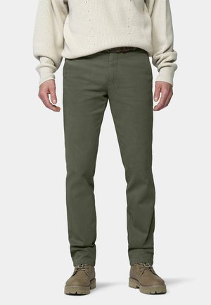 Mann trägt olivgrüne Hose, beigen Strickpullover und braune Schnürstiefel, steht vor einem schlichten Hintergrund.