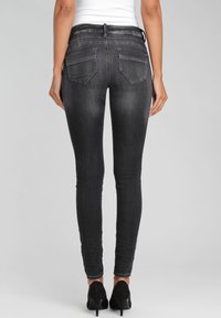 Los pantalones vaqueros de corte ajustado en negro presentan dos bolsillos traseros, un acabado texturizado y una cintura de altura media. Llevados con tacones altos negros.