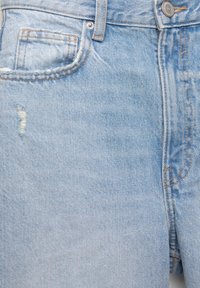 Jean en denim bleu clair avec une petite déchirure usée près de la poche avant et une fermeture par bouton métallique à la taille.