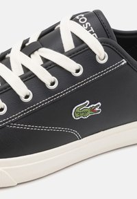 Baskets en cuir noir avec lacets blancs, œillets argentés, coutures blanches et un logo crocodile vert brodé sur le côté.