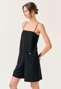 Zwarte jumpsuit met spaghettibandjes, met een gestructureerde lijfje, zijzakken met gouden accenten en een losvallend kort ontwerp.