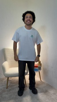 Mann med krøllete hår som smiler, iført hvit Adidas-t-skjorte, svarte bukser og svarte sko, står ved siden av hvit teksturert lenestol og et sidebord med bøker og vase.