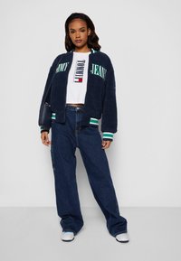 Tumesinise fleece bomber jakk roheliste ja valgete triipudega, millele on sobitatud valge lühike T-särk logoga ja laiad tumedad teksapüksid.