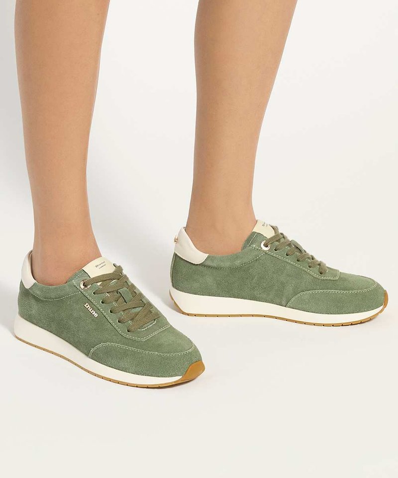 Des pieds portant des baskets en daim vert à lacets, semelles blanches et détails beiges sur un fond clair uni.