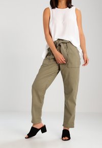 Kvinna som bär en ärmlös vit blus, högmidjade olivgröna byxor med tygbälte, och svarta klackskor med öppen tå, stående på vit golv.