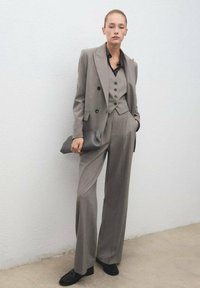Traje gris de tres piezas que presenta una chaqueta cruzada, un chaleco ajustado y pantalones de pierna ancha. Combinado con un elegante clutch negro y zapatos.
