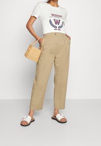 Witte katoenen t-shirt met de opdruk "WEEKEND", beige hoge taillebroek en witte sandalen met zilveren accenten. Geweven beige handtas.