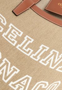 Sac fourre-tout en tissu tissé beige avec texte souligné de blanc et poignées en cuir marron avec coutures blanches.