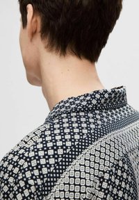 Camicia a maniche corte con sfondo navy e motivi geometrici bianchi, colletto aperto e tessuto liscio e leggero.