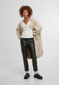 Beige oversize kardigan vyrobený z texturované tkaniny, nošený přes bílý fitted top s detailem uzlu, spárovaný s černými koženými kalhotami a mokasínami.